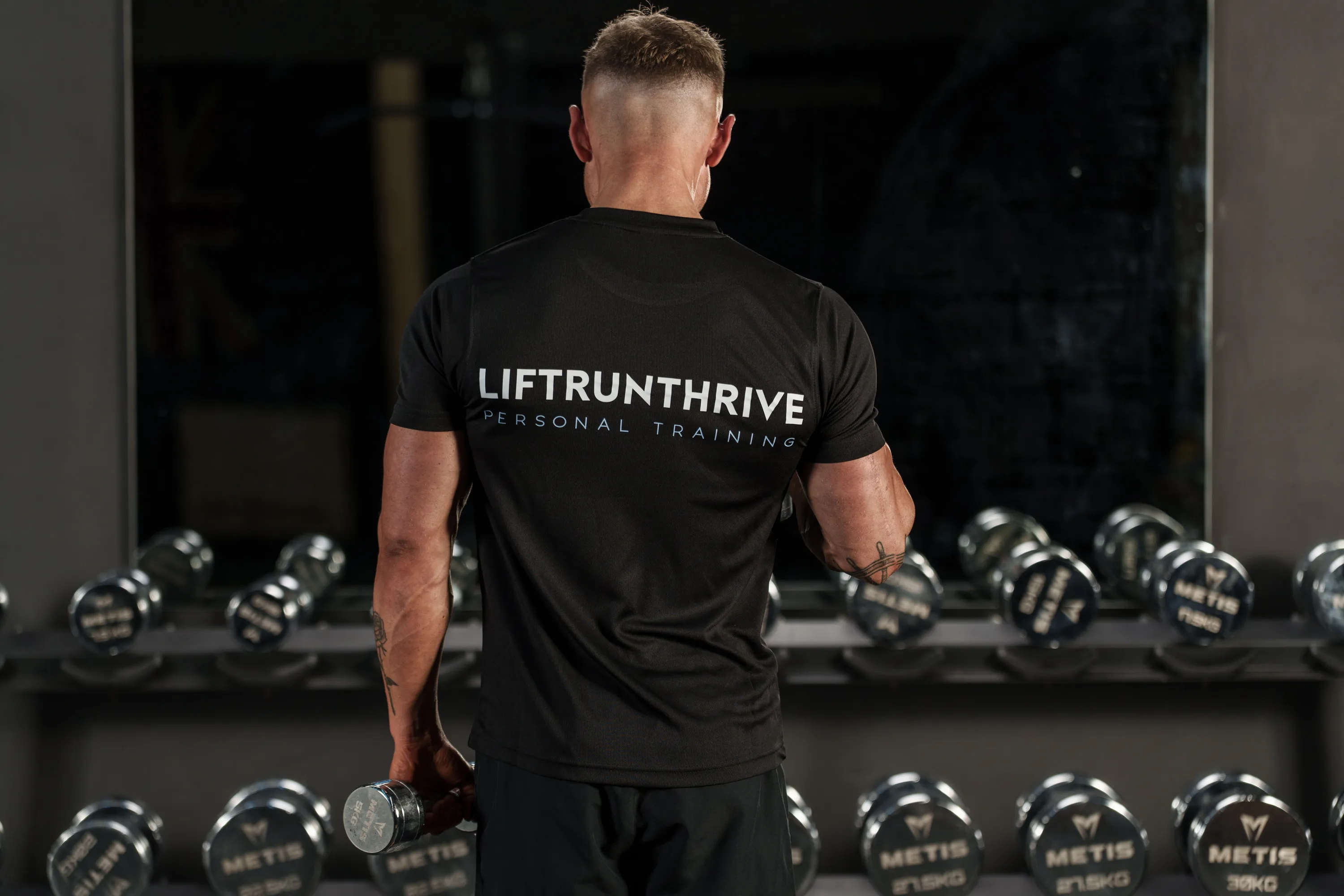 Callum from behind in a Lift Run Thrive personal training t-shirt, facing a dumbbell rack